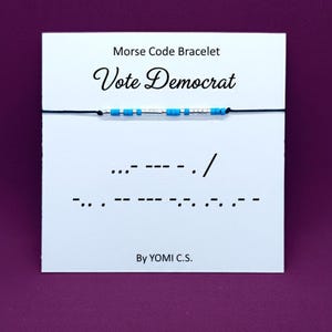 May include: A Morse code bracelet with blue and silver beads spelling "Vote Democrat" on a white card. The card also features Morse code symbols and the text "By YOMI C.S."