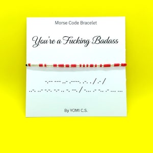 May include: A Morse code bracelet with red beads and gold accents is displayed on a white card. The card reads "You're a Fucking Badass" in cursive, with Morse code symbols below. The card is on a bright yellow background.