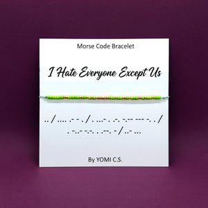 May include: A Morse code bracelet with the message "I Hate Everyone Except Us" on a white card. The bracelet features green and gold beads. The card also includes the Morse code translation and the text "By YOMI C.S."