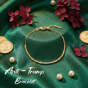 May include: A gold-colored bracelet with a delicate chain and cylindrical beads. The bracelet is displayed on a green satin surface, adorned with pearls, coins, and burgundy flowers. The text "Anti-Trump Bracelet" is visible.