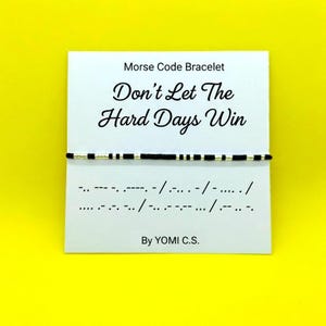 May include: A Morse code bracelet featuring alternating black and silver beads on a white display card. The card displays the text "Morse Code Bracelet", "Don't Let The Hard Days Win", the corresponding Morse code sequence, and "By YOMI C.S.".