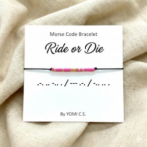May include: A Morse code bracelet featuring pink and gold beads on a black cord, spelling "Ride or Die". The bracelet is presented on a white card with the text "Morse Code Bracelet" and "By YOMI C.S."
