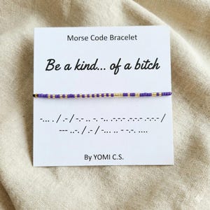 May include: A Morse code bracelet with purple and gold beads is displayed on a white card. The card reads "Be a kind... of a bitch" in black script. Below the text is the Morse code translation. The card is signed "By YOMI C.S."
