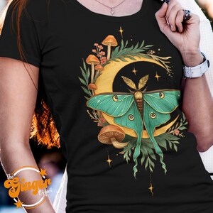 May include: Black t-shirt featuring a teal moth with gold accents, set against a gold crescent moon and mushrooms. The design includes botanical elements and stars, creating a whimsical, nature-inspired aesthetic.