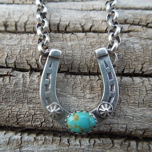May include: A silver horseshoe pendant necklace with a turquoise stone in the center. The horseshoe is decorated with small silver squares and stars.