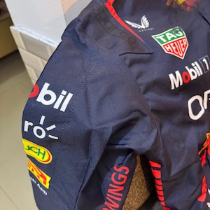 May include: Navy blue racing jacket with sponsor logos. Features the "Mobil 1" and "TAG Heuer" logos, along with "Oracle" and "Red Bull Racing" branding. The jacket has red and yellow accents and the words "YOU WINGS" on the sleeve.