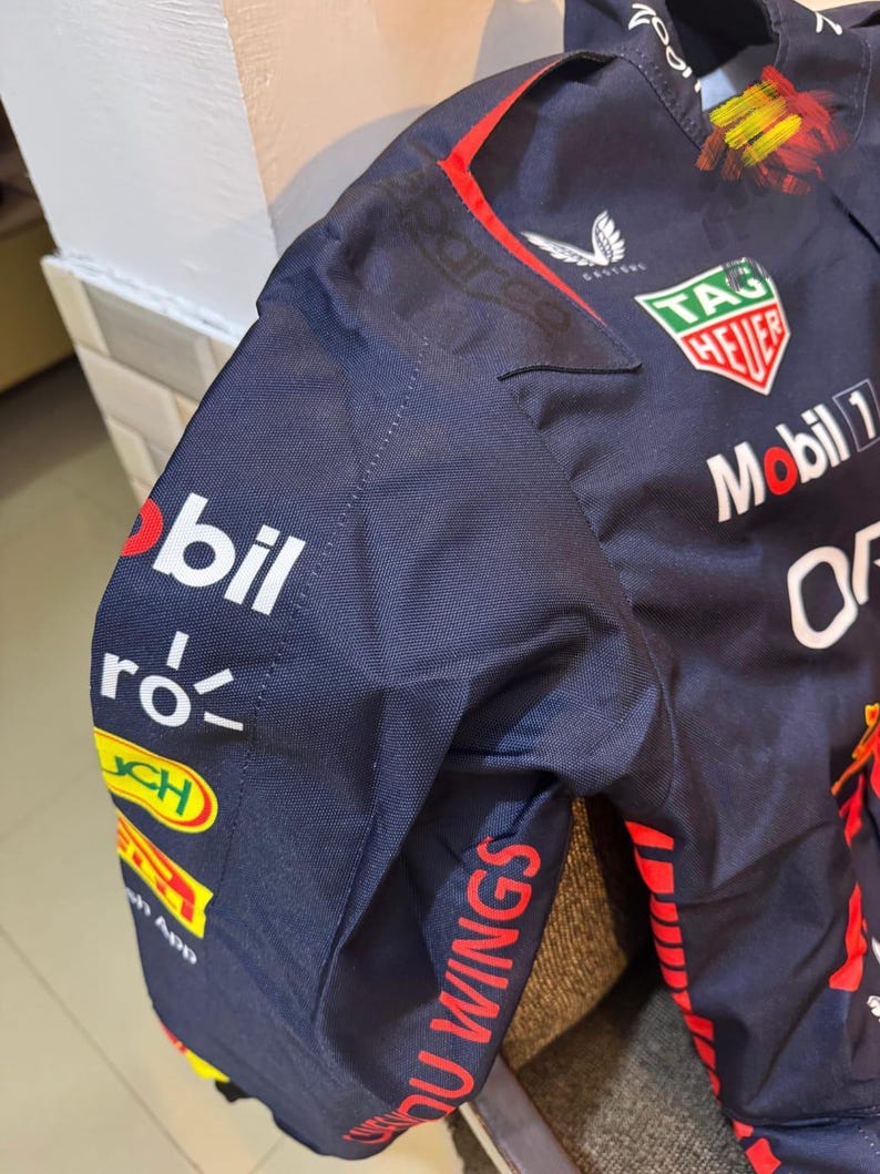 May include: Navy blue racing jacket with sponsor logos. Features the "Mobil 1" and "TAG Heuer" logos, along with "Oracle" and "Red Bull Racing" branding. The jacket has red and yellow accents and the words "YOU WINGS" on the sleeve.
