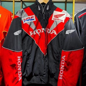 Puede incluir: Chaqueta de carreras Honda negra, roja y gris con el logotipo de Honda. La chaqueta tiene cremallera completa, cuello alzado y el logotipo HRC. Las mangas son negras con detalles rojos y la palabra "HONDA" en blanco.