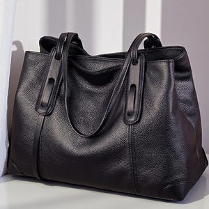 Könnte beinhalten: Eine schwarze Ledertasche mit doppelten Henkeln und einem strukturierten Design. Die Tasche hat eine genarbte Textur und steht auf einer weißen Oberfläche. Die Griffe haben ein einzigartiges Design mit einem Metallakzent.