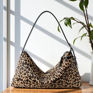 Puede incluir: Bolso de hombro con estampado de leopardo, correa y cremallera negras. El bolso está hecho de un material suave y texturizado y descansa sobre una superficie de madera. El bolso tiene forma triangular.
