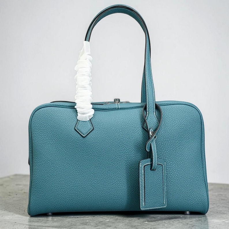 May include: Teal-colored leather handbag with a rectangular shape and a top handle. The bag features a textured surface, a silver-tone zipper, and a matching luggage tag. The handle is wrapped in white protective material.