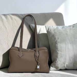 May include: A taupe-colored leather handbag with long, slender handles. The rectangular bag features a silver-toned zipper and a small lock detail. The bag is set against a neutral-toned background with soft pillows.