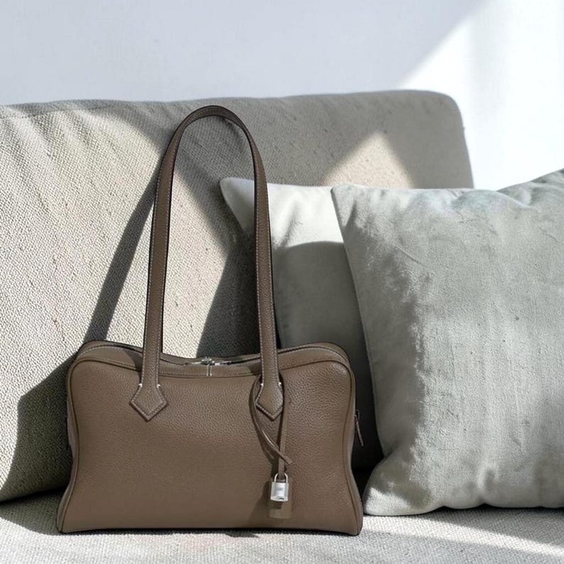 May include: A taupe-colored leather handbag with long, slender handles. The rectangular bag features a silver-toned zipper and a small lock detail. The bag is set against a neutral-toned background with soft pillows.