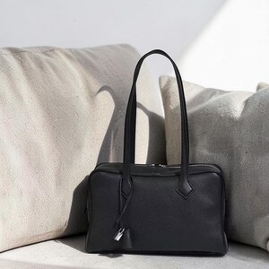 May include: A black leather handbag with long handles, a rectangular shape, and a small silver-colored lock charm. The bag is set against a backdrop of neutral-toned pillows, highlighting its sleek design and texture.