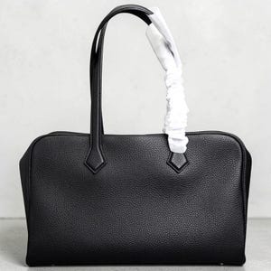 May include: A black leather handbag with a rectangular shape and two top handles. The bag has a textured surface and diamond-shaped accents near the handles. A white protective wrapping is attached to one handle.
