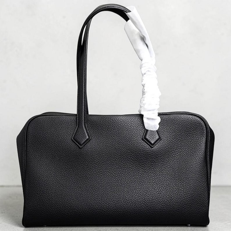 May include: A black leather handbag with a rectangular shape and two top handles. The bag has a textured surface and diamond-shaped accents near the handles. A white protective wrapping is attached to one handle.