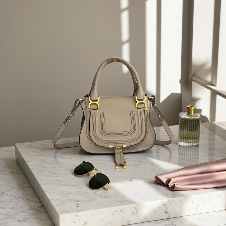 May include: A light gray handbag with a top handle and a shoulder strap, featuring gold-colored hardware. The bag is paired with black sunglasses, a perfume bottle, and a pink scarf. The items are arranged on a marble surface.