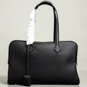 May include: A black leather tote bag with a rectangular shape and two top handles. The bag features a textured surface, a zipper closure, and a decorative leather tag. A white fabric is wrapped around one of the handles.