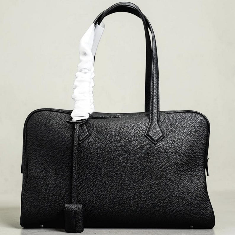 May include: A black leather tote bag with a rectangular shape and two top handles. The bag features a textured surface, a zipper closure, and a decorative leather tag. A white fabric is wrapped around one of the handles.