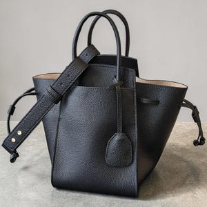 May include: Black leather tote bag with a structured design. Features a top handle, a detachable shoulder strap, and a drawstring closure. The bag has a textured exterior and a light-colored interior lining. Includes a small, attached leather tag.