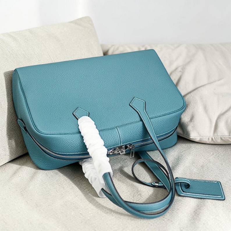 May include: A turquoise leather handbag with a rectangular shape and a zipper closure. The bag has two top handles and a detachable shoulder strap. A white protective wrap covers the handles. A matching luggage tag is attached to the strap.