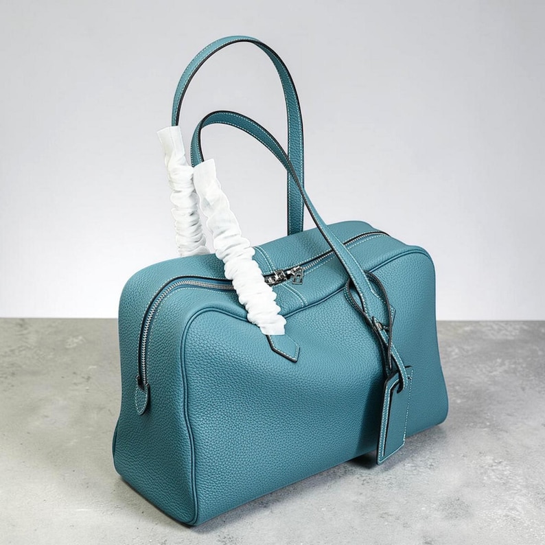 May include: A teal-colored leather handbag with a rectangular shape and dual handles. The bag features a zipper closure, a small tag, and white protective wrapping on the handles. The stitching is white, and the bag has a textured surface.