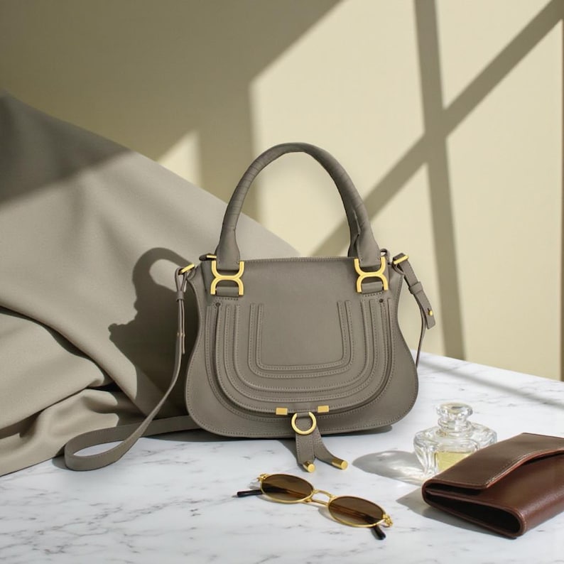 May include: A gray leather handbag with a top handle and gold-colored hardware. The bag has a curved shape with decorative stitching. A pair of gold-rimmed sunglasses, a brown wallet, and a perfume bottle are also present.