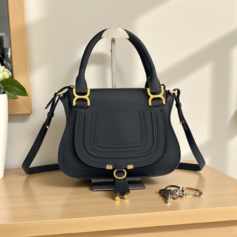 May include: Black leather handbag with gold-tone hardware. The bag features a curved design with a top handle and a detachable shoulder strap. The front has a decorative stitched detail and a gold ring with a tassel.