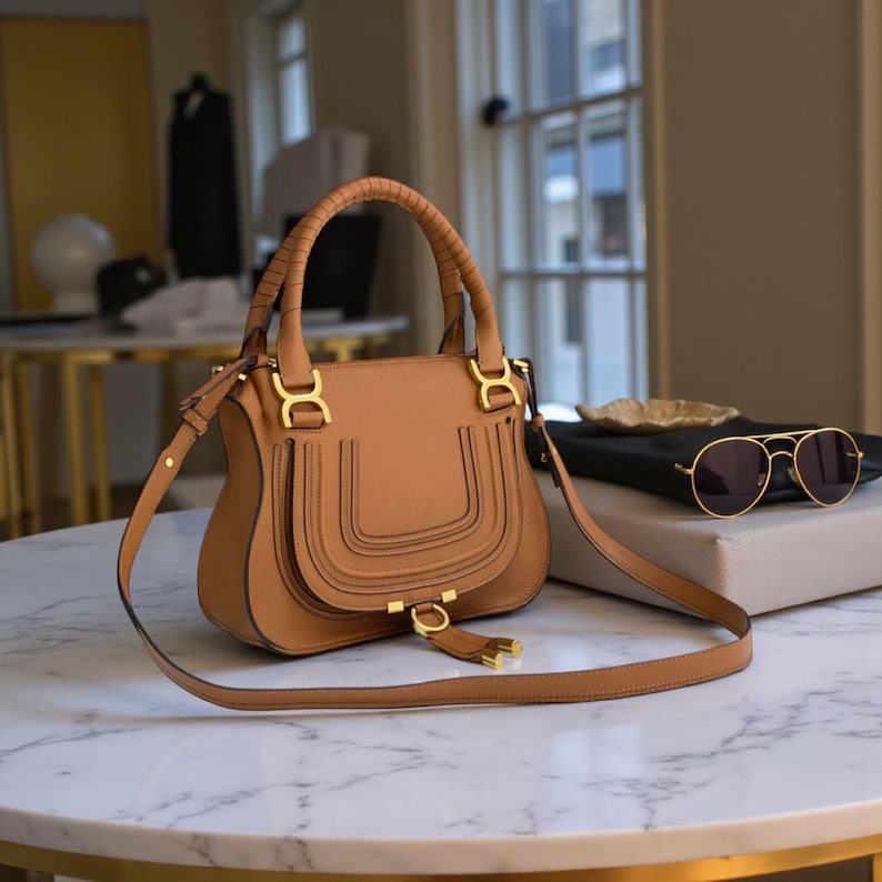 May include: A tan leather handbag with a top handle and a detachable shoulder strap. The bag features gold-colored hardware and decorative stitching. A pair of sunglasses rests on a nearby surface.