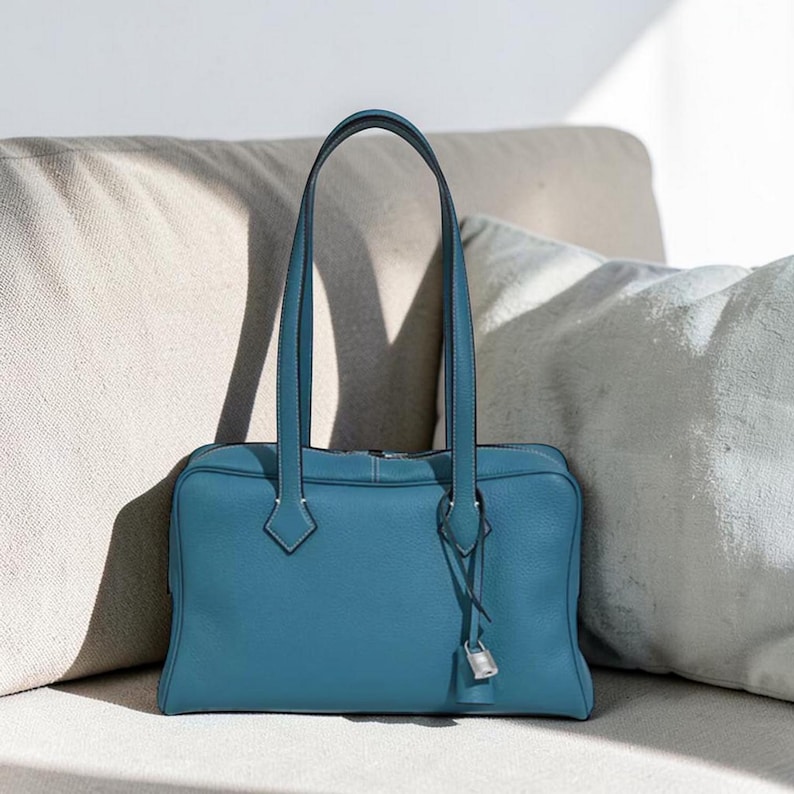 May include: A teal leather handbag with a rectangular shape and two long handles. The bag features a silver-colored lock and key detail. The bag is sitting on a beige couch with white pillows.