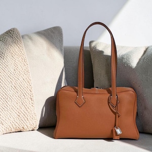 May include: A rectangular, tan leather handbag with two long handles. The bag has a silver-colored lock and key charm hanging from the side. The bag is sitting in front of two neutral-colored pillows.