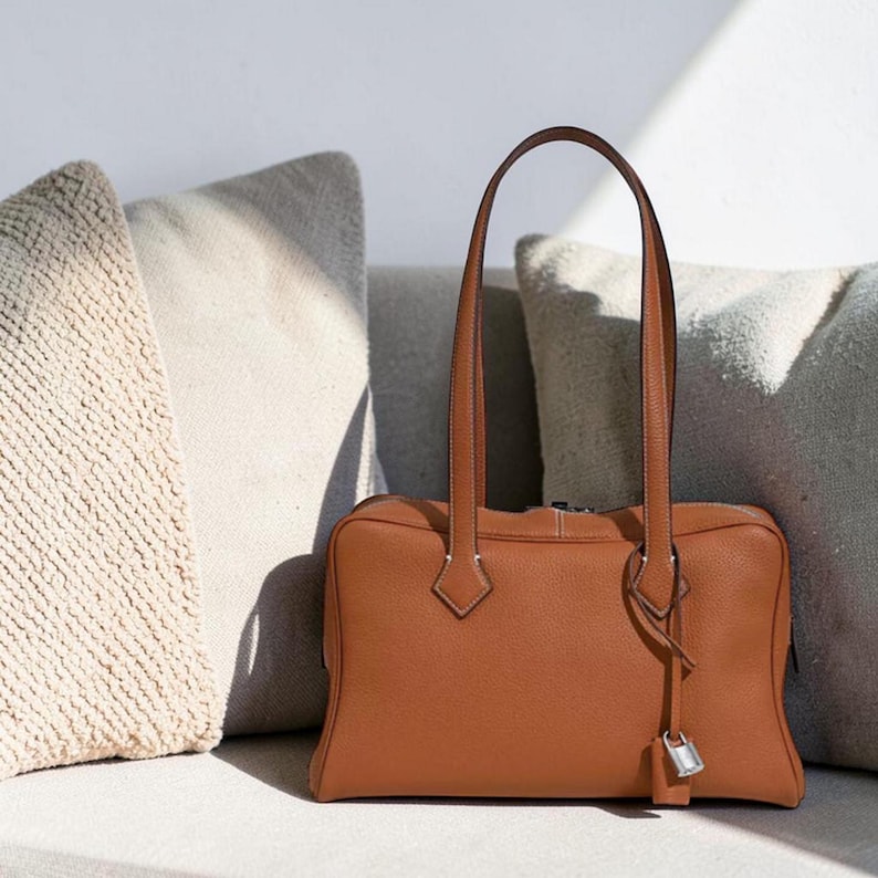 May include: A rectangular, tan leather handbag with two long handles. The bag has a silver-colored lock and key charm hanging from the side. The bag is sitting in front of two neutral-colored pillows.