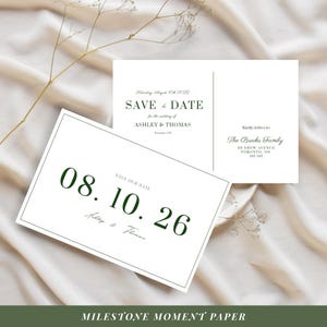 May include: White wedding stationery with dark green text. One card reads "Save the Date" for Ashley & Thomas, with the date August 10th, 2026. Another card displays the date "08.10.26". The cards are on a cream-colored fabric.