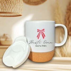 Könnte beinhalten: Weiße Keramik-Tasse mit einer rosa Schleifen-Grafik und den Worten "Beste Oma" und "LILLY & THEA" in schwarzer Schrift. Der Boden der Tasse ist aus Kork, und ein weißer Deckel liegt daneben. Die Tasse hat einen Henkel.