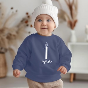 Może przedstawiać: Niemowlę w granatowej bluzie i białej dzianinowej czapce. Bluza ma białą grafikę świecy z cyfrą "1" i słowem "one". Dziecko się uśmiecha. Idealne na pierwsze urodziny.
