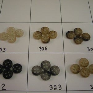 May include: Assortment of round buttons in various colors, including light tan, brown, black, and gray. Each button has four holes and is arranged in groups. The numbers 303, 306, 307, 312, 323, and 304 are visible.