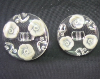 May include: Three clear, round buttons with a floral design. Each button features off-white rose-shaped embellishments and silver-colored swirls. The buttons vary in size, with the largest measuring approximately 1.25 inches in diameter.