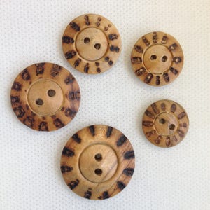 May include: A collection of five wooden buttons in varying sizes. Each button has two holes and a light brown center. The buttons are decorated with dark brown, irregular patterns around the edges, giving them a rustic appearance.