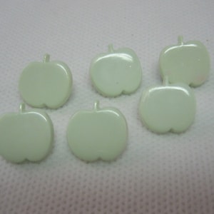 Puede incluir: Seis botones en forma de manzana verde pálido. Cada botón tiene un tallo y una superficie lisa y brillante. Los botones están dispuestos sobre una superficie blanca.