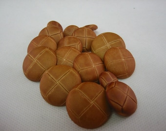 May include: A collection of brown, round buttons with a cross-stitch design. The buttons are made of a leather-like material and have a slightly textured surface. The buttons are arranged in a loose pile against a white background.