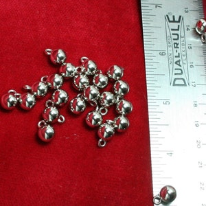 May include: A collection of small, shiny, silver-colored ball charms with attached loops, scattered on a red velvet surface. A ruler with inch measurements is in the background. These charms could be used for jewelry making or other crafts.