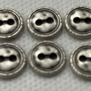 May include: Six antique silver-colored buttons with two holes each. The buttons have a slightly raised, textured rim and a matte finish. These buttons are suitable for sewing projects and garment embellishment.