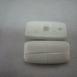 May include: Two white, rectangular plastic components. One component has a small, raised rectangular feature and several small holes. The other component has a segmented design. Both components are on a light gray surface.