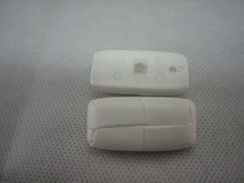 May include: Two white, rectangular plastic components. One component has a small, raised rectangular feature and several small holes. The other component has a segmented design. Both components are on a light gray surface.