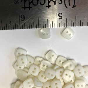 May include: A collection of off-white, square buttons with two holes each. A metal ruler is placed over the buttons. The buttons are approximately 0.4 inches in size, suitable for various sewing projects.