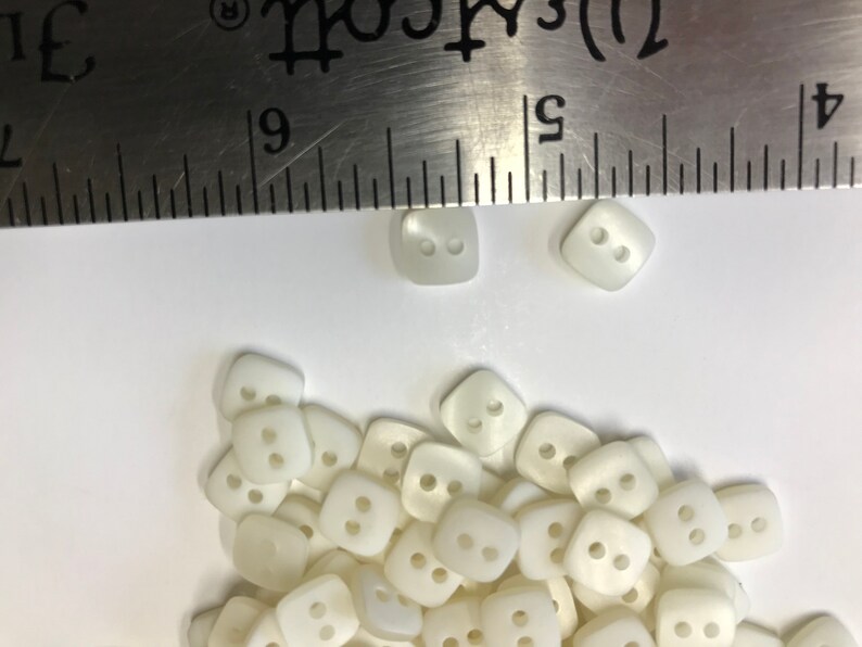 May include: A collection of off-white, square buttons with two holes each. A metal ruler is placed over the buttons. The buttons are approximately 0.4 inches in size, suitable for various sewing projects.