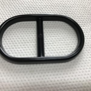May include: A black oval-shaped belt buckle with a vertical bar in the center. The buckle is made of a smooth, solid material and is set against a white, textured background. The design is simple and modern.
