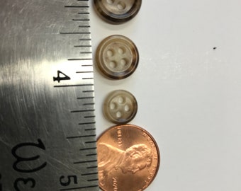 Puede incluir: Tres botones marrones con cuatro agujeros cada uno, de diferentes tamaños, se muestran junto a una regla y un centavo. Los botones tienen un aspecto veteado. El botón más grande tiene aproximadamente 1,9 cm de diámetro.