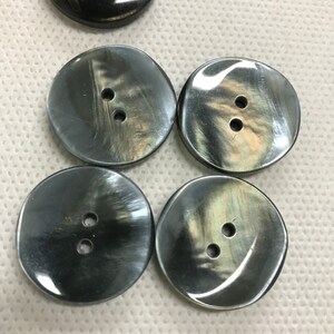 K&ouml;nnte beinhalten: Eine Sammlung runder, dunkelgrauer Kn&ouml;pfe mit jeweils zwei L&ouml;chern. Die Kn&ouml;pfe haben einen perlmuttartigen Glanz, der das Licht reflektiert. Ein schwarzer Knopf ist ebenfalls sichtbar. Diese Kn&ouml;pfe eignen sich f&uuml;r N&auml;h- und Bastelprojekte.