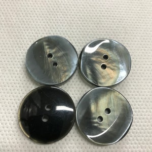 Puede incluir: Cuatro botones redondos e iridiscentes con dos agujeros cada uno. Los botones son una mezcla de negro y gris, con un acabado brillante. Están dispuestos en un patrón cuadrado, adecuados para coser o manualidades.